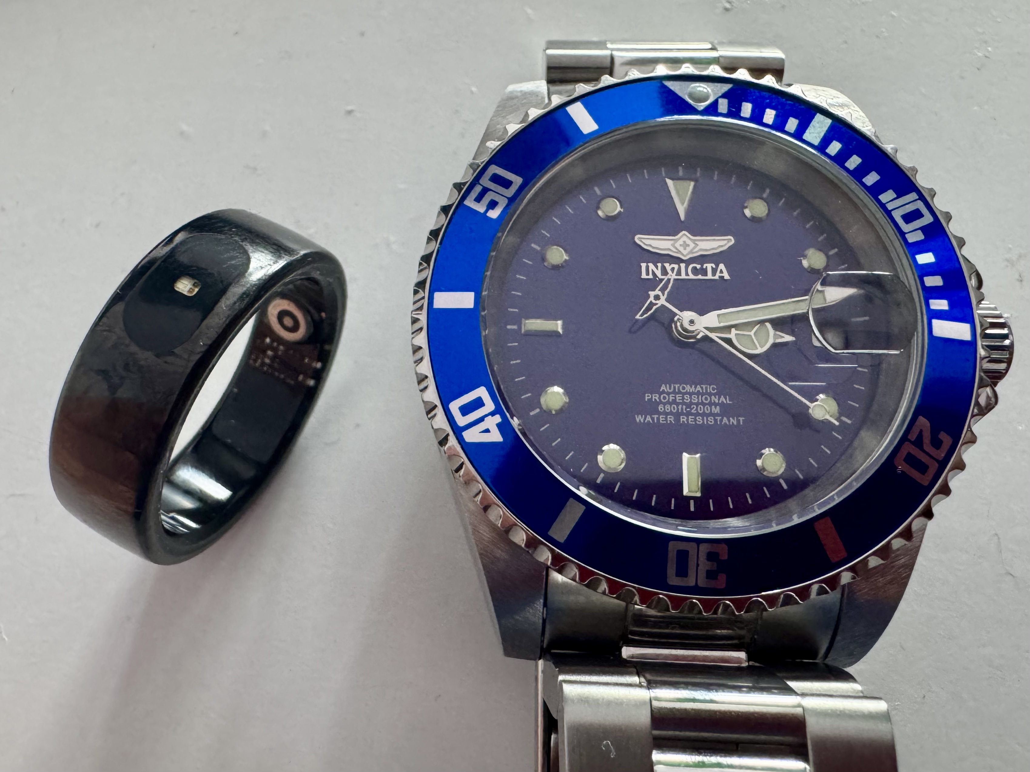 zegarek i smart ring Czarny smart ring znajduje się obok niebieskiego automatycznego profesjonalnego zegarka Invicta z niebieską ramką.