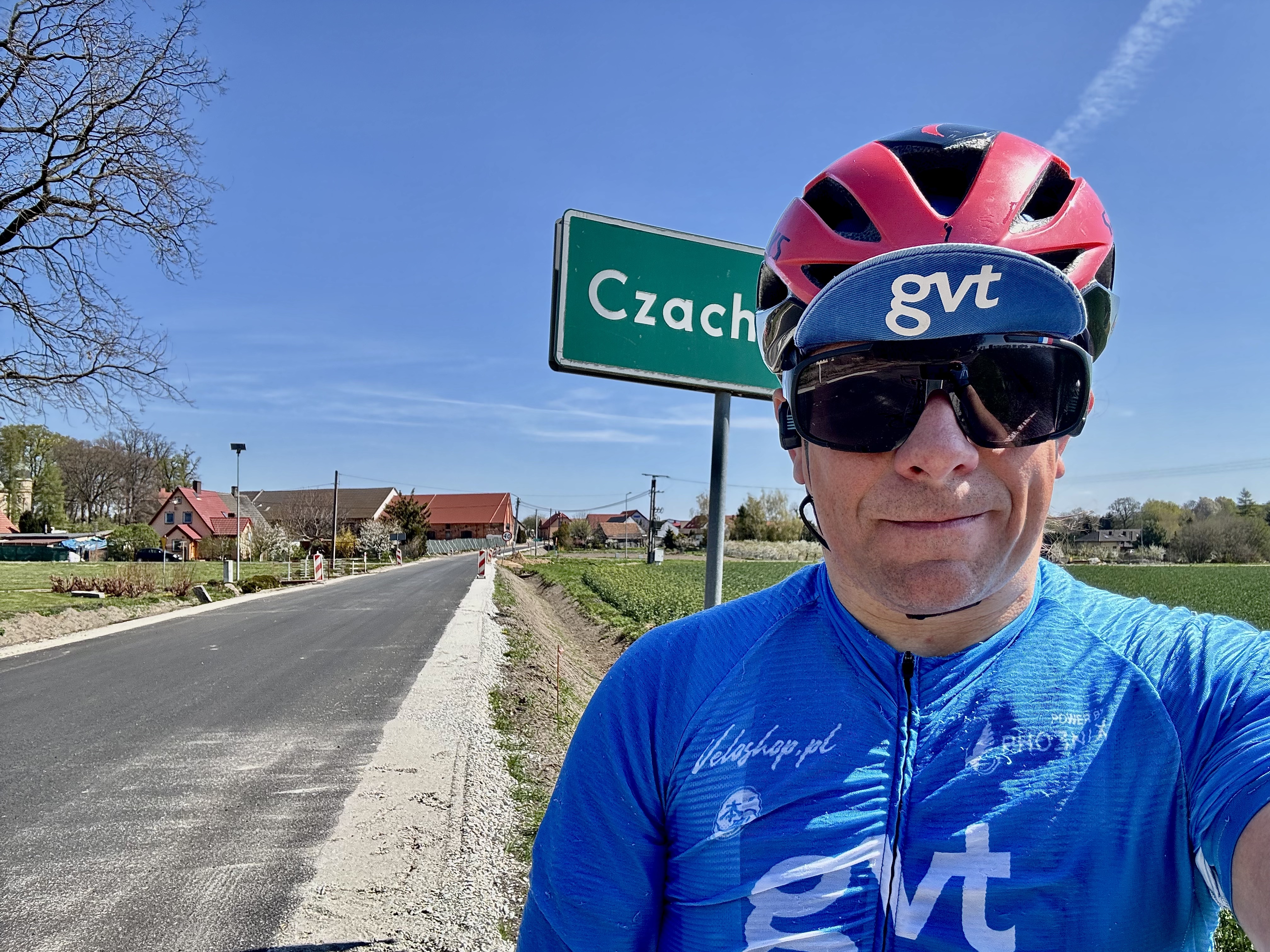 Rowerzysta w niebieskiej koszulce i czerwonym kasku w okularach przeciwsłonecznych stoi w pobliżu znaku z napisem „Czach” na wiejskiej drodze z domami i polami pod czystym błękitnym niebem.
