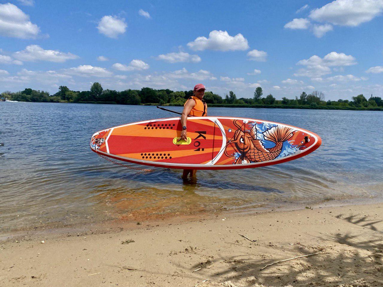 Osoba ubrana w kamizelkę ratunkową trzyma kolorową deskę SUP z motywem ryby koi stojąc w płytkiej wodzie na piaszczystej plaży w słoneczny dzień.